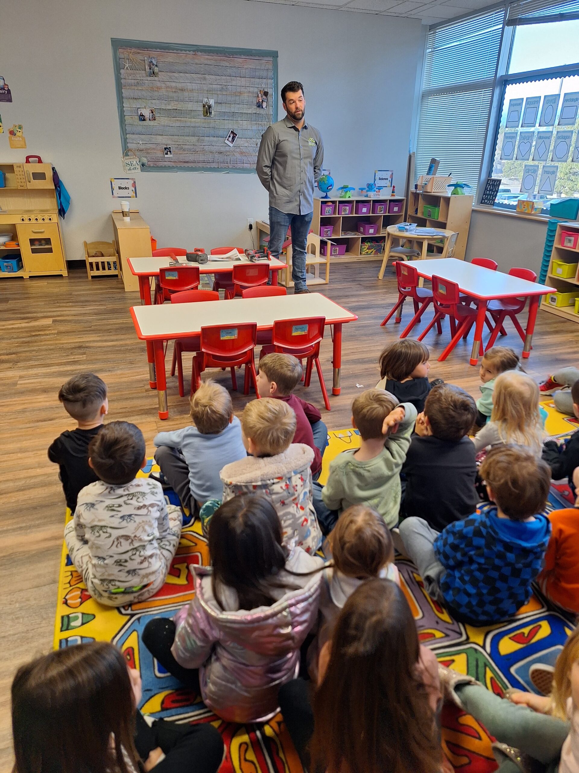 A teacher stands at the front of a colorful classroom, reminiscent of creative commercial construction, engaging with attentive children. The room features small tables and chairs, educational posters on the walls, and toys on shelves as the children sit attentively on a patterned rug.