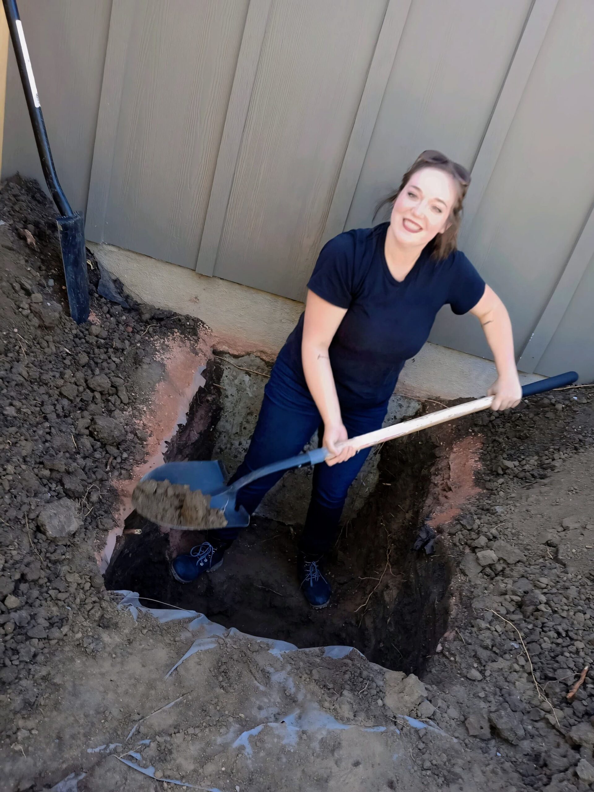 A woman in a black shirt and jeans is smiling while shoveling dirt in a deep hole next to a building, embodying the dedication synonymous with NewEra Construction. Another shovel leans against the wall in the background.