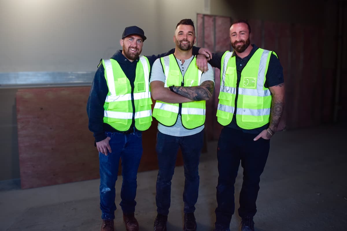 Three men wearing neon yellow safety vests stand together in a commercial construction site. They are smiling and have a casual appearance, with one man having his arms around the others. The background shows the unfinished walls of their ongoing project.