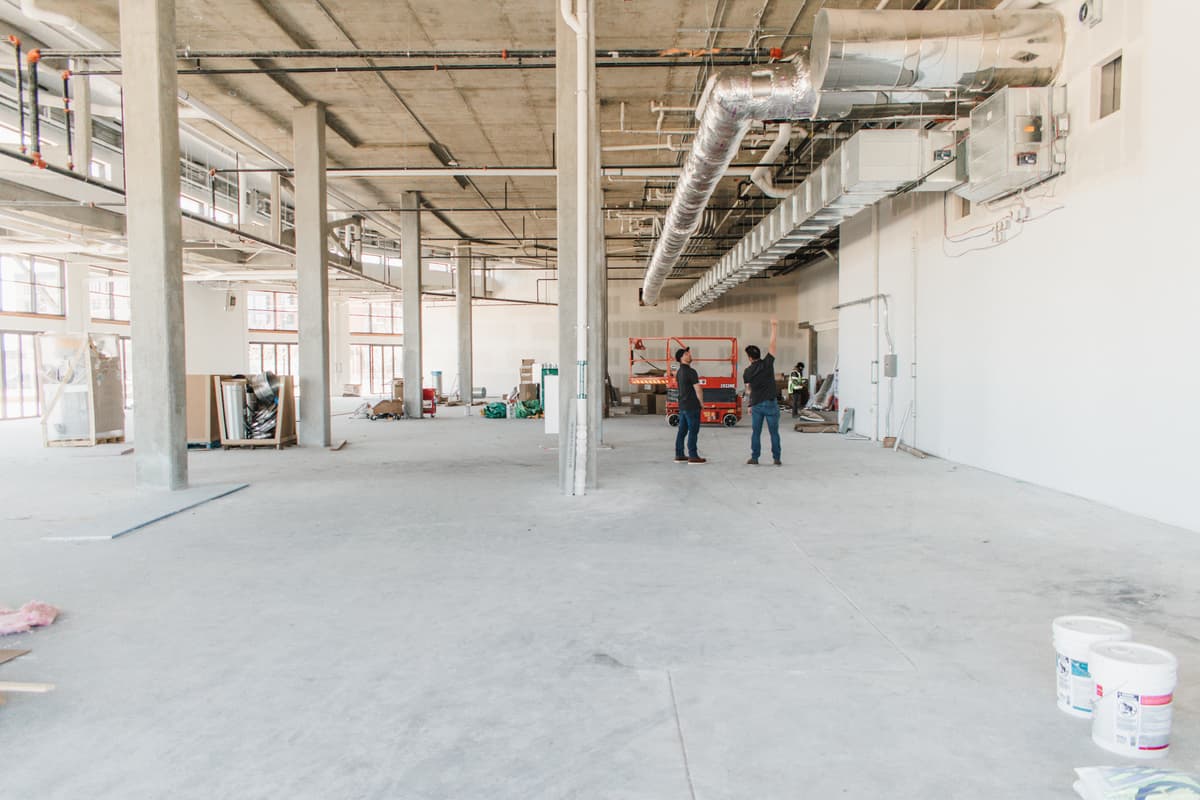 A large, unfinished industrial space with concrete floors, exposed ducts, and support pillars hints at a commercial construction project. Two people stand near the center, surrounded by construction materials and tools. Natural light streams through large windows, highlighting ongoing renovation work.