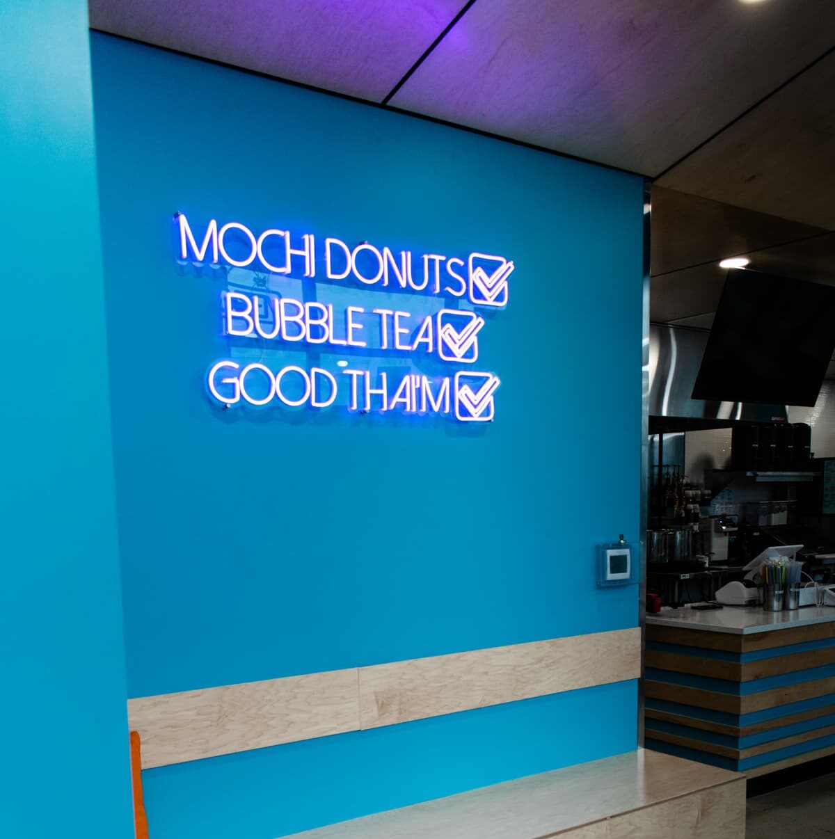 A brightly lit cafe wall, showcasing skilled commercial construction, features a neon sign with three enticing lines: "Mochi Donuts," "Bubble Tea," and "Good Thaim," each accented with a checkmark. The blue backdrop is complemented by a wooden bench beneath the vibrant display.