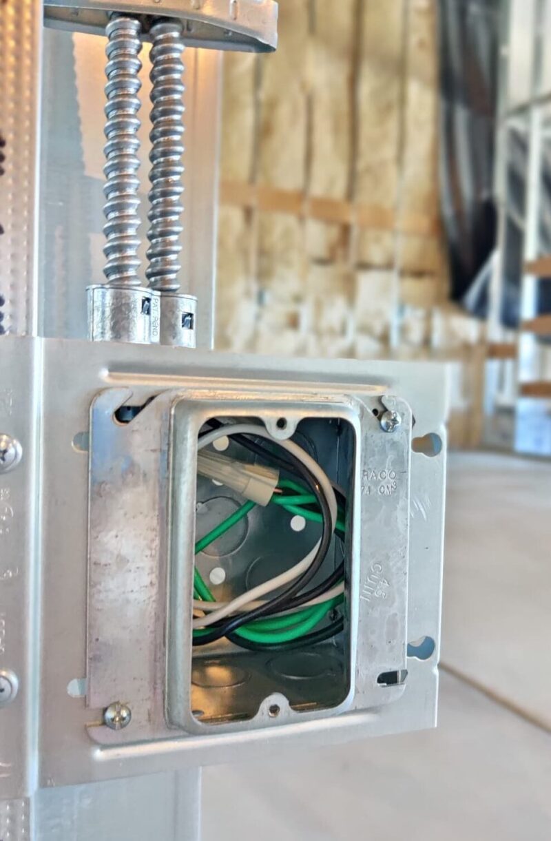 A partially installed electrical outlet box with exposed wiring is mounted on a metal frame, characteristic of commercial construction projects. Green, white, and black wires are visible inside the box against a backdrop of unfinished construction materials and insulation.