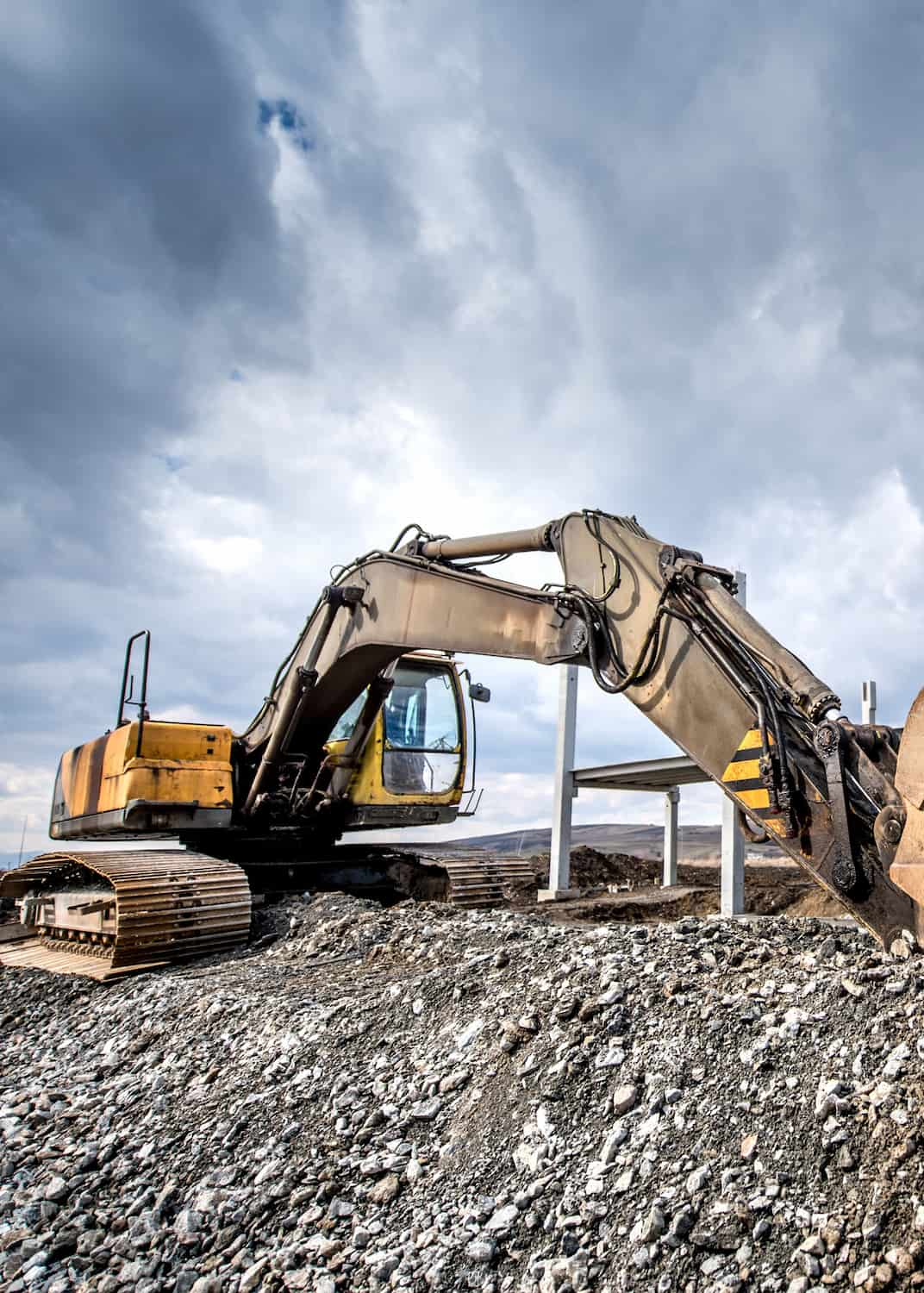 A large excavator with a yellow and brown color scheme sits on a rocky commercial construction site under a cloudy sky. The machine's arm extends forward, resting on the uneven terrain.