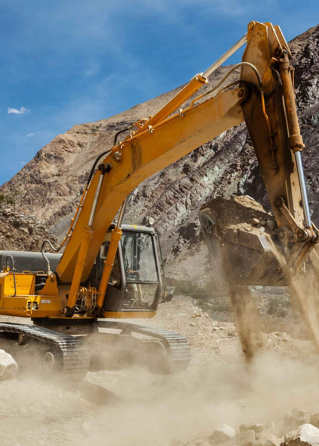 A yellow excavator from NewEra Construction is digging into the ground, lifting dirt and rocks in a mountainous area. The sky is clear and blue, with dust visible around the machine.