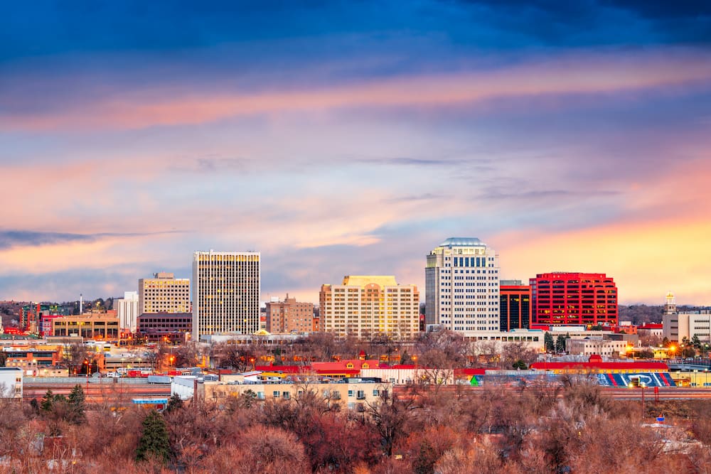 A city skyline at sunset showcases a mix of modern, commercial construction and mid-rise buildings. The sky is painted with soft pastel hues of pink, orange, and blue. In the foreground, leafless trees are visible, silhouetted against the evening light.