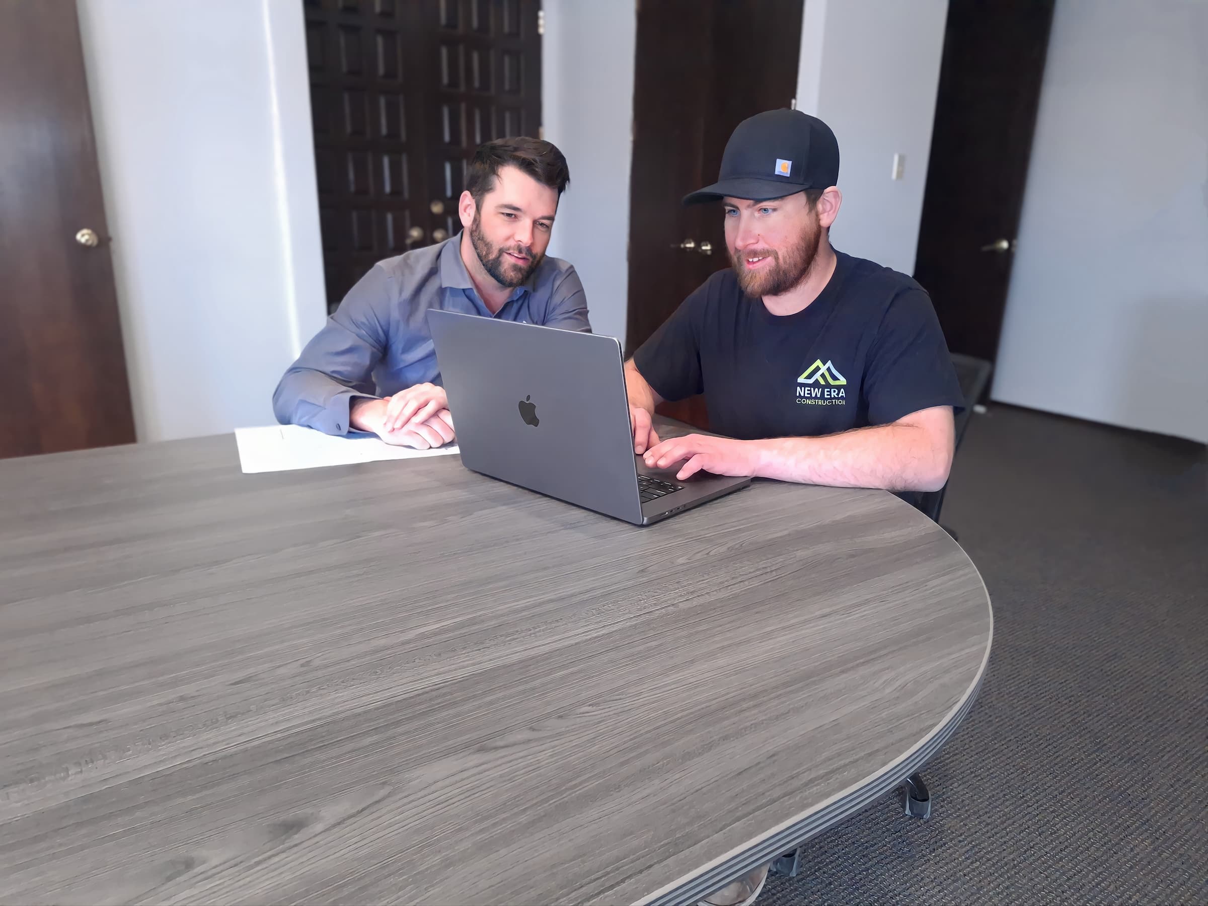 Two men sit at a table in an office setting, focused on a laptop. One wears a gray shirt, the other a black shirt and cap. They seem to be discussing commercial construction plans on the screen. A document lies beside the laptop, detailing the project.