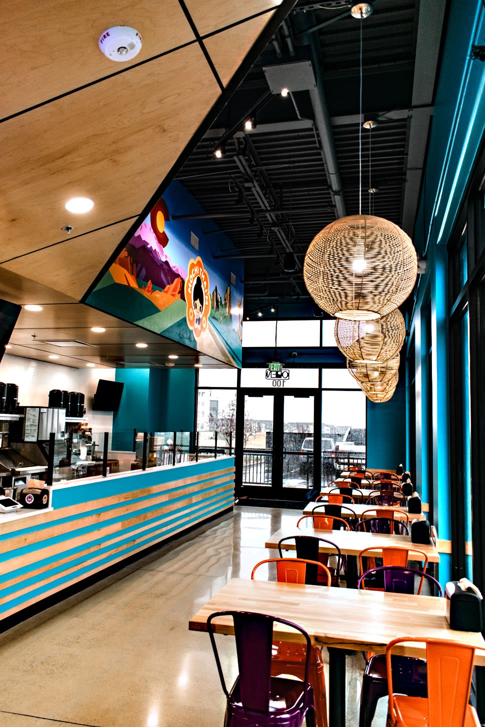 A modern, vibrant cafe interior created in collaboration with a skilled commercial general contractor. The colorful decor includes wood panel ceilings and turquoise walls. Orange and purple chairs surround the tables under large light fixtures, while a desert landscape mural adds charm to this inviting space.