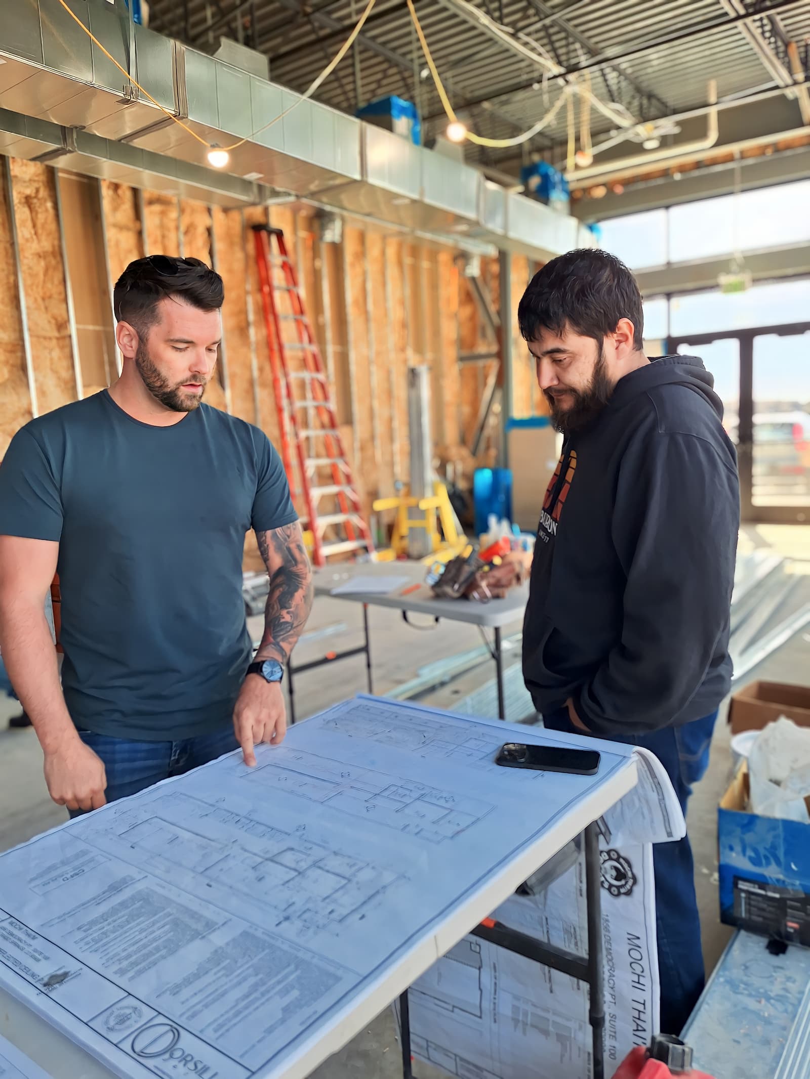 Two men are standing in a partially constructed building, discussing a set of blueprints on a table. The room, indicative of commercial construction, has exposed wooden beams and visible wiring. A red ladder and various construction materials are in the background.