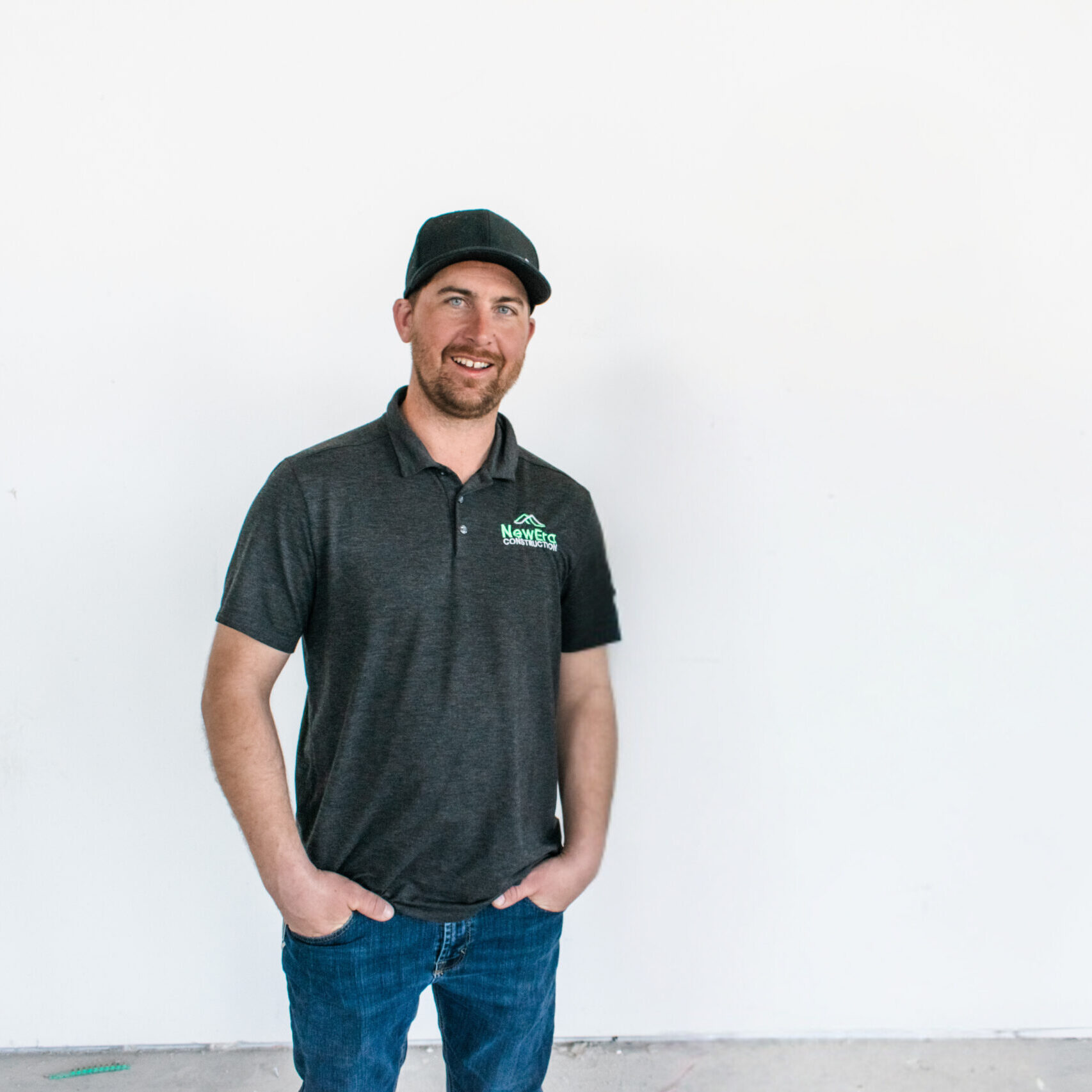 A man in a dark polo shirt and jeans stands against a plain white background. Sporting a black NewEra Construction cap, he has his hands in his pockets, smiling confidently at the camera.