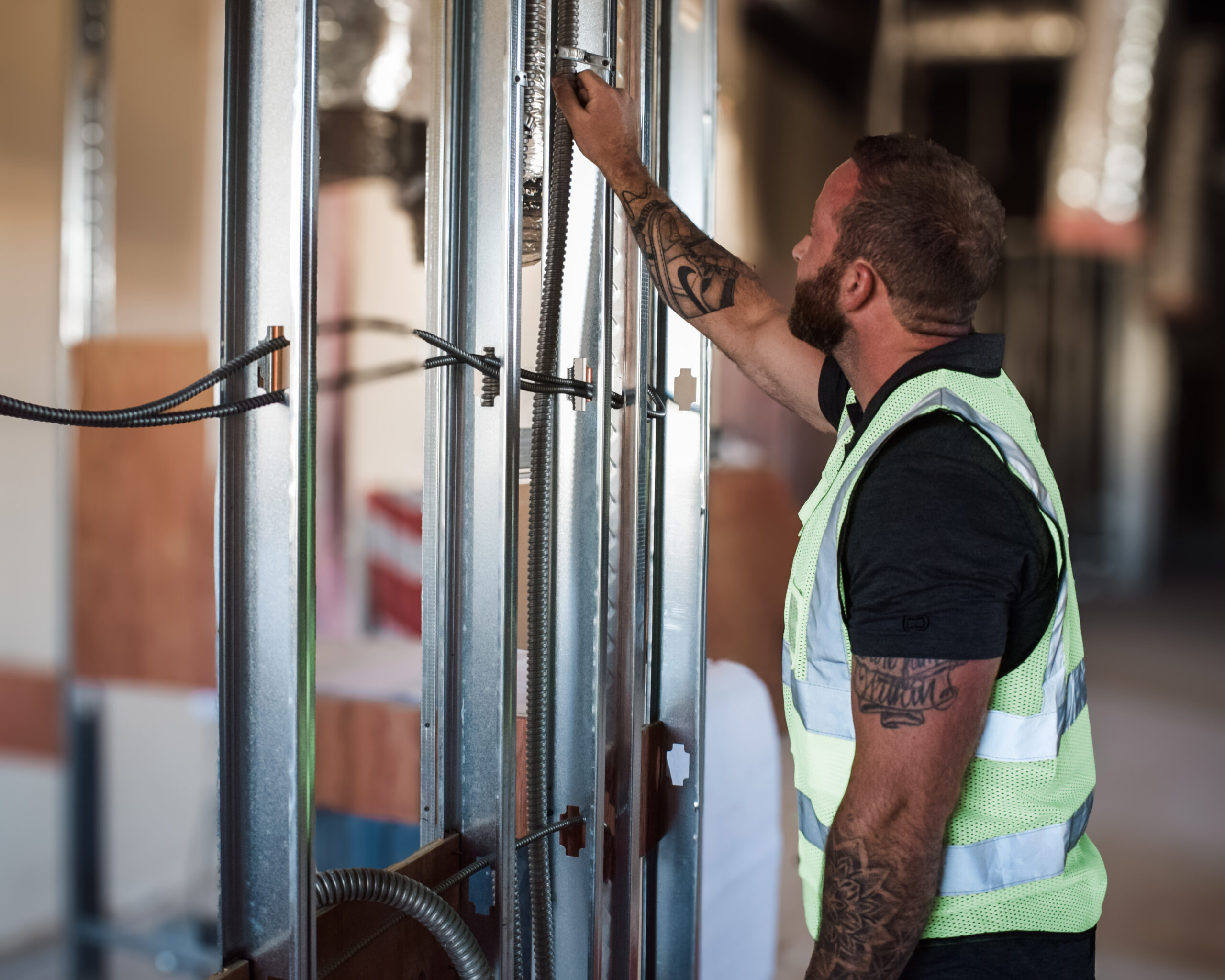 A construction worker, sporting a reflective vest and black shirt, diligently installs electrical cables within metal wall studs. This scene is part of a larger commercial construction services project, featuring exposed framing in the building's unfinished setting.