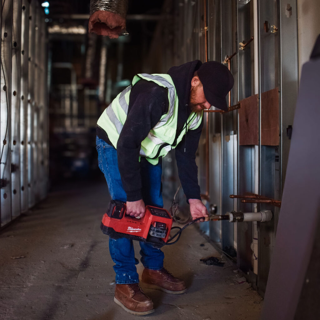 A construction worker, donning a safety vest and cap, operates a power tool against a metal wall frame inside a building under construction. This industrial setting—marked by exposed pipes and insulation—showcases the precision and expertise synonymous with NewEra Construction.