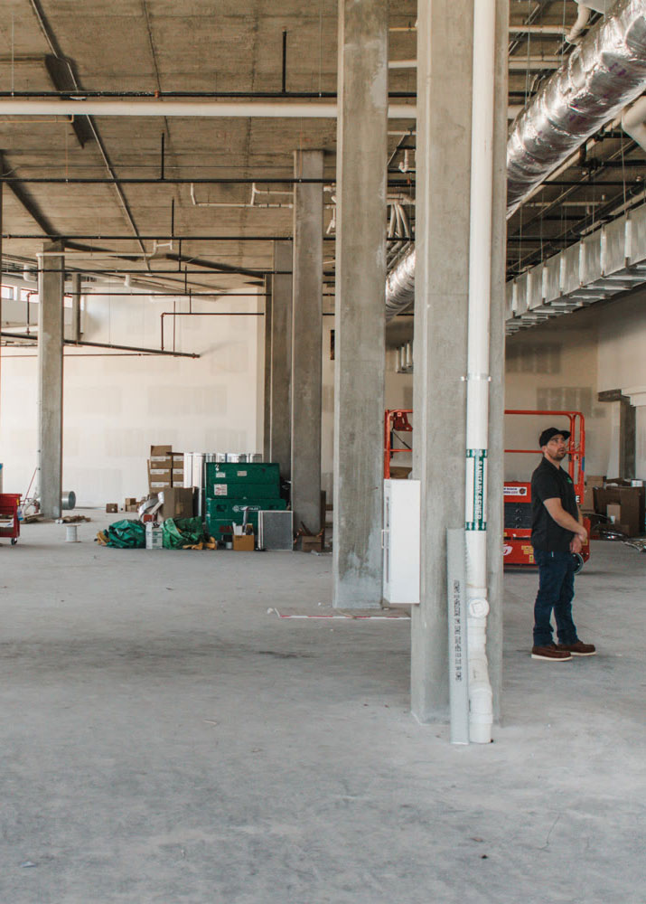 A man stands inside an unfinished building with concrete floors and exposed ceilings. As a commercial general contractor, he navigates through boxes, construction materials, and equipment scattered around the space. Large pipes are visible, and a green dumpster looms in the background.