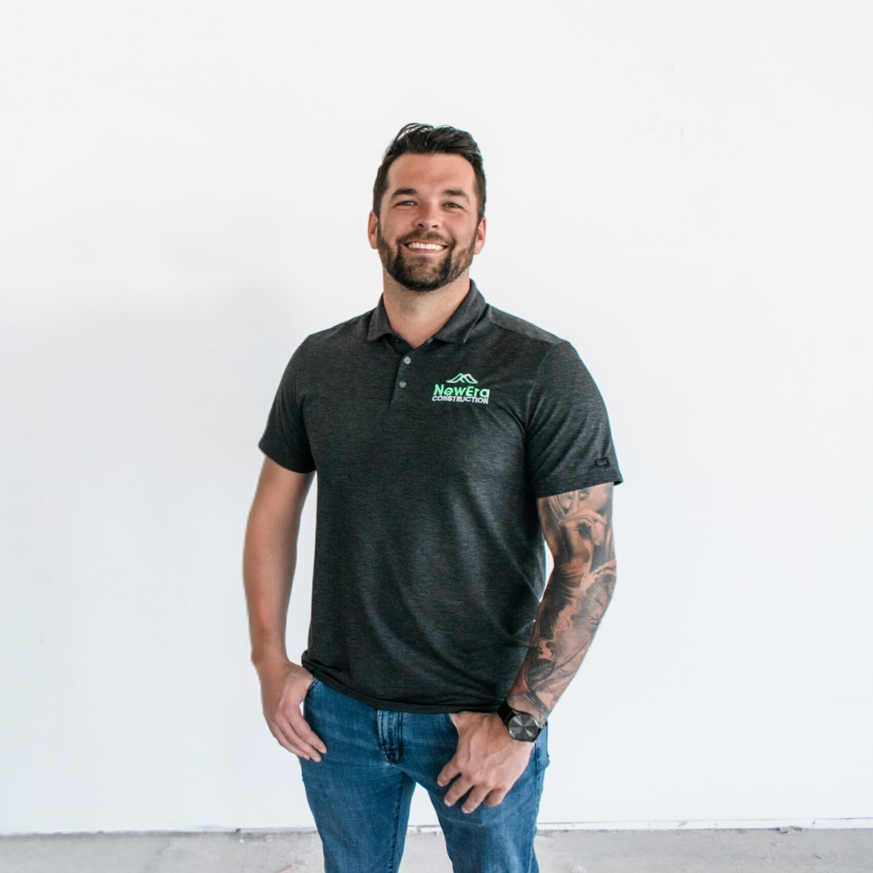 A man with short dark hair and a beard stands smiling, wearing a dark polo shirt with a NewEra Construction logo on the left side and blue jeans. He has a sleeve tattoo on his left arm and stands against a plain white background.