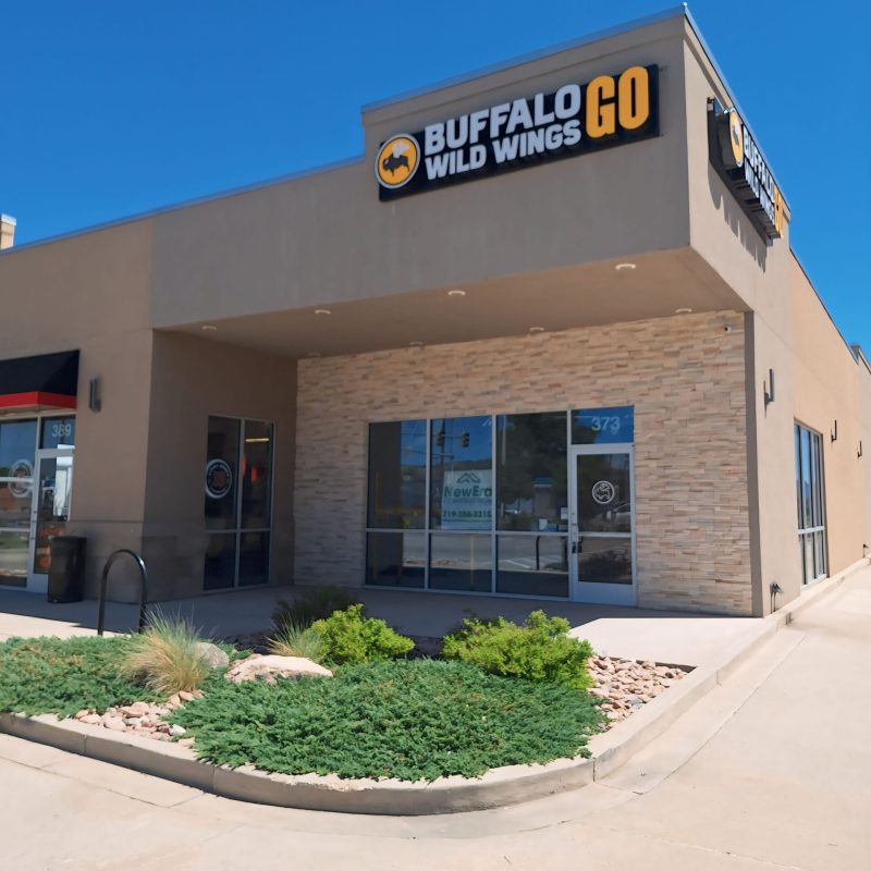 The exterior of the Buffalo Wild Wings GO restaurant showcases a stunning example of commercial construction, featuring a beige facade and large windows. The logo is prominently displayed above an entrance framed by lush greenery under a clear, blue sky.