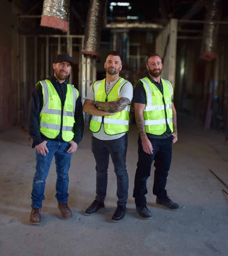 Three construction workers from NewEra Construction, wearing yellow high-visibility vests, stand in a partially constructed building. The floor is unfinished, surrounded by various materials. They smile confidently at the camera, embodying the spirit of NewEra Construction's commitment to excellence.