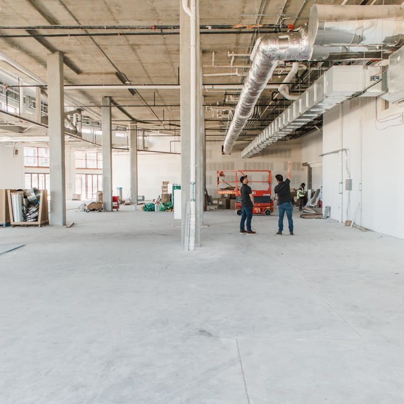 A large, unfinished industrial space with concrete floors, exposed ducts, and support pillars hints at a commercial construction project. Two people stand near the center, surrounded by construction materials and tools. Natural light streams through large windows, highlighting ongoing renovation work.