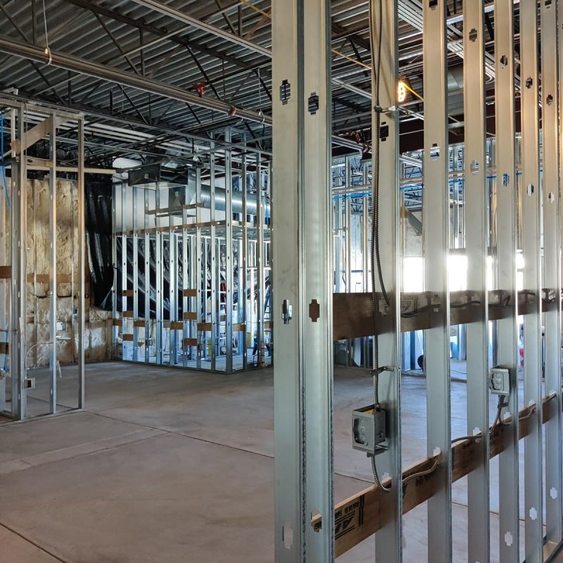 A commercial construction site reveals metal wall studs set on a concrete floor. The unfinished interior displays exposed beams and electrical boxes, with insulation visible on some walls. Sunlight streams through an opening, illuminating the evolving space.