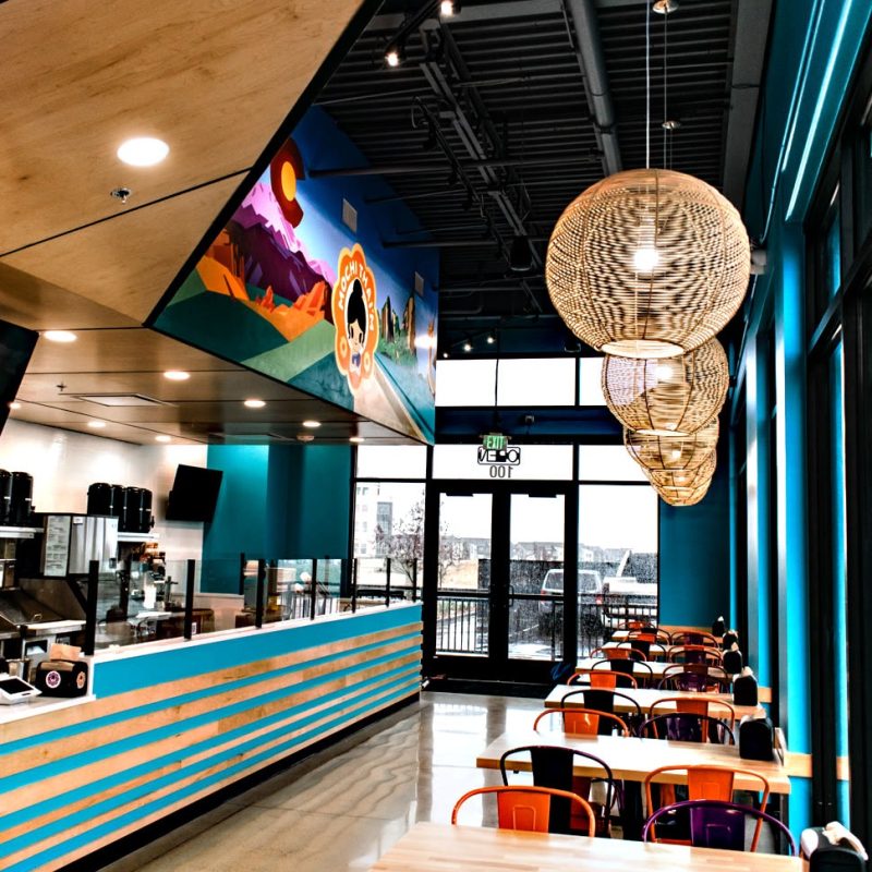 A modern, vibrant cafe interior created in collaboration with a skilled commercial general contractor. The colorful decor includes wood panel ceilings and turquoise walls. Orange and purple chairs surround the tables under large light fixtures, while a desert landscape mural adds charm to this inviting space.