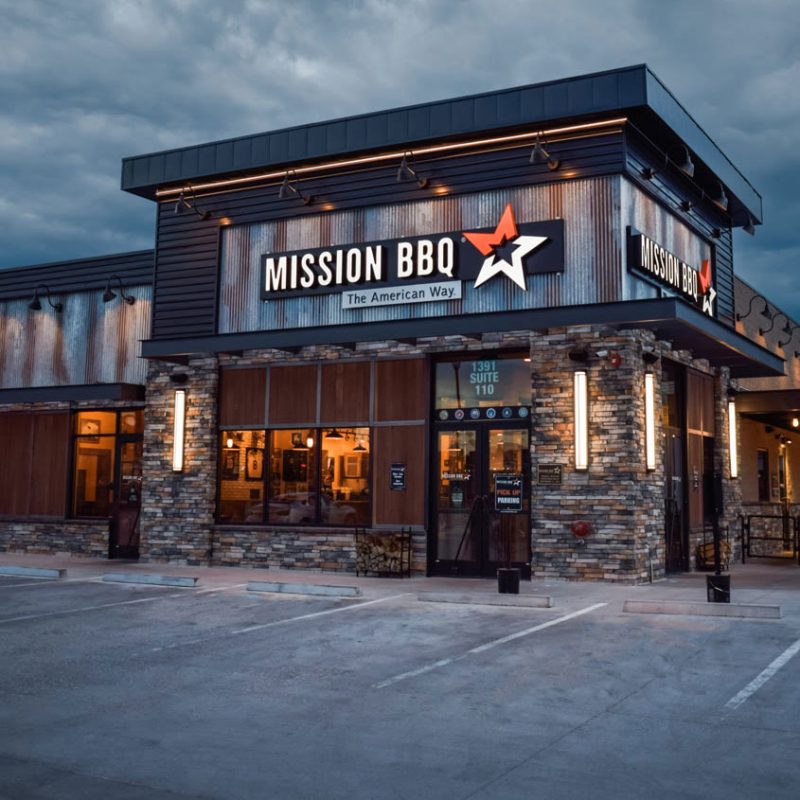 The exterior of a Mission BBQ restaurant, crafted by a skilled commercial general contractor, showcases rustic stone and wood design. The logo features a star with the sign "The American Way." Amidst a cloudy sky, the well-lit building exudes a warm and inviting atmosphere.