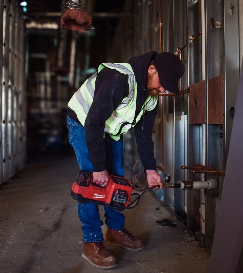 A construction worker, donning a safety vest and cap, operates a power tool against a metal wall frame inside a building under construction. This industrial setting—marked by exposed pipes and insulation—showcases the precision and expertise synonymous with NewEra Construction.