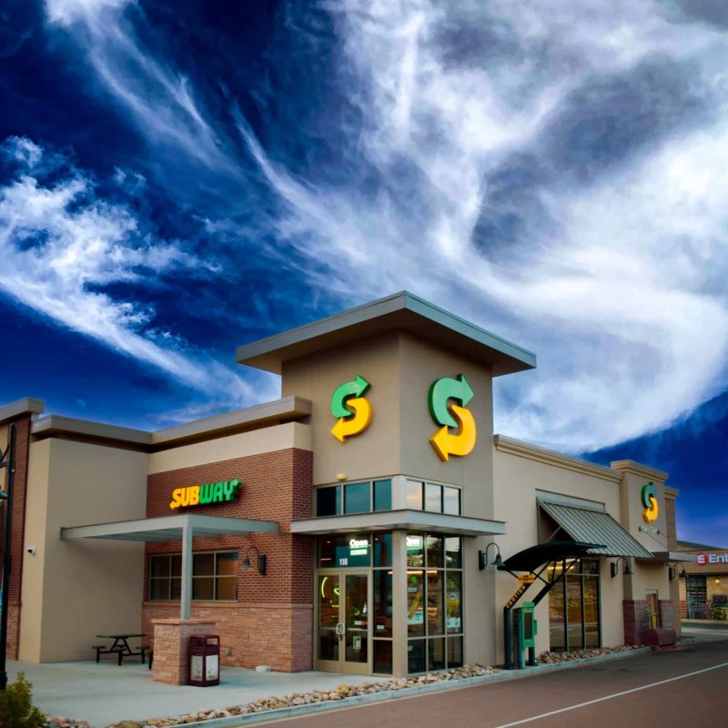 A modern Subway restaurant, brought to life by a skilled commercial general contractor, features a tan facade with large windows and its distinctive green and yellow logo. Under a vibrant blue sky with wispy clouds, you'll find a bicycle rack and outdoor seating in front.